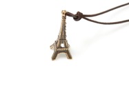 Gustowny wisiorek wykonany z metalu nieszlachetnego w kolorze starego złota, miniatura paryskiej wieży Eiffla, zawieszony na brązowym skórzanym rzemyku